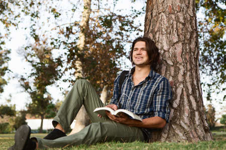 Young man sitting against tree holding book, looking away smilingの写真素材