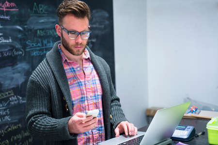 Young man in workplace using laptop looking at smartphoneの写真素材