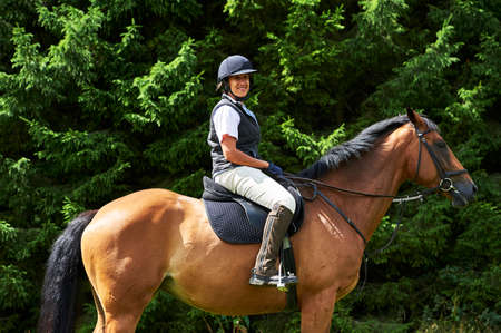 Side view of mature woman on horseback wearing riding hat and boots looking at camera smiling