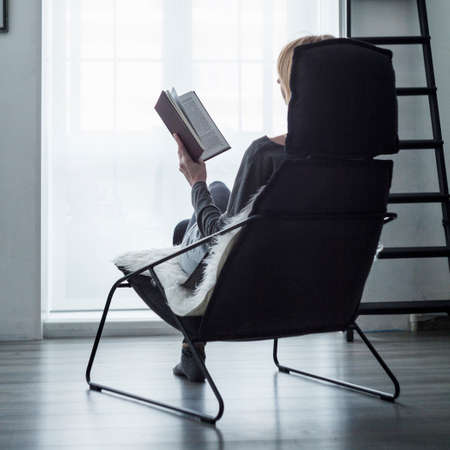 Woman sitting on chair in front of window reading a bookの写真素材