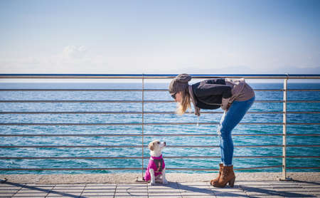 Full length side view of young woman by railings in front of ocean bending over looking at dogの写真素材