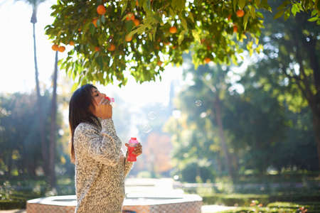 Side view of mature woman blowing bubbles under orange tree, Seville, Spainの写真素材