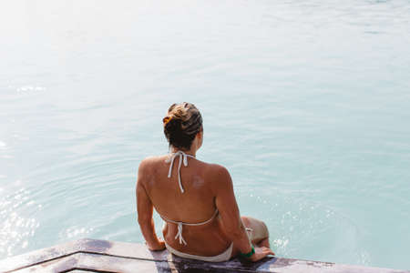 High angle rear view of mid adult woman wearing bikini sitting on pier, Blue Lagoon hot springs, Icelandの写真素材