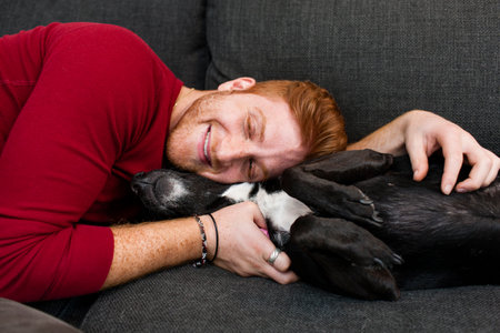 Young man lying on sofa hugging pet dog eyes closed smilingの写真素材