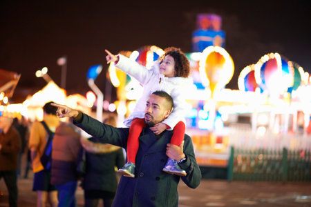 Father carrying daughter on shoulders at funfair, father and daughter pointing