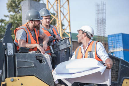 Site manager talking with steamroller drivers about blueprint on construction site