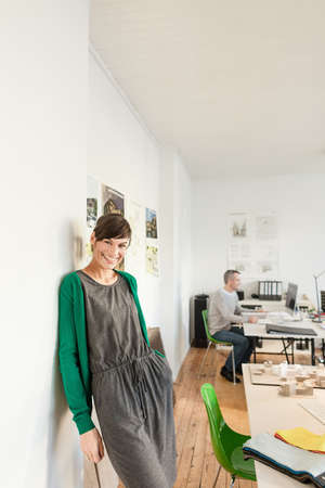 Mature woman in office leaning against wall looking at camera smiling