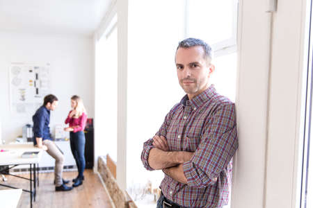 Mature man leaning against window arms crossed looking at camera smilingの写真素材