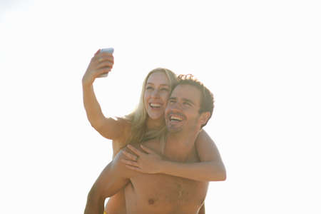 Man giving woman piggyback using smartphone to take selfie, smilingの写真素材