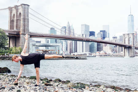 Mid adult man practicing yoga on riverside in front of Brooklyn Bridge, New York, USA