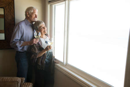Senior couple looking out of window, holding glass of wine, smilingの写真素材