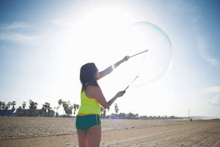 Rear view of woman on beach, arms raised using bubbles wands to make bubblesの写真素材