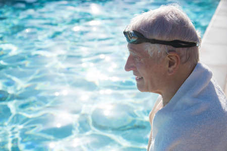Senior man sitting on edge of outdoor swimming pool, towel around shoulders