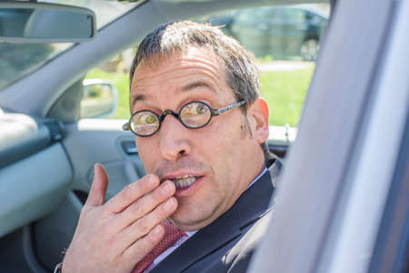 Man in car wearing eye glasses, hand over mouth looking at camera