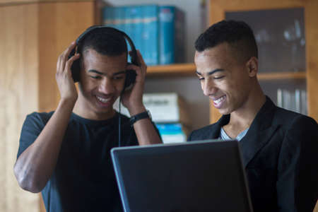 Twin brothers at home, looking at laptop, wearing headphones, smilingの写真素材
