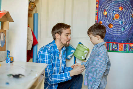 Father helping son button shirt in bedroom