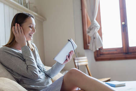 Woman sitting on bed wearing headphone looking at digital tablet smiling