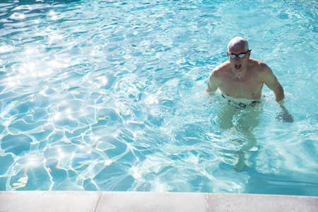 Senior man emerging from water in outdoor swimming poolの写真素材