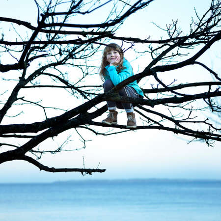 Girl in tree by ocean sitting on branch, hand on chin looking awayの写真素材