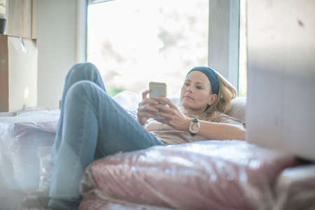 Moving house: woman relaxing on plastic wrapped sofa, using smartphone