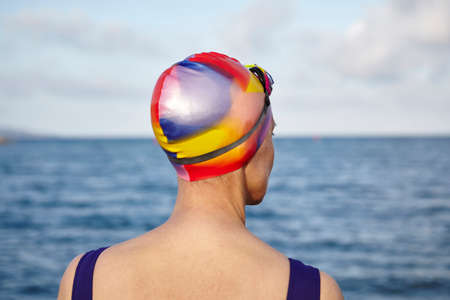 Mature woman wearing swimming costume and swimming hat, standing beside the sea, rear viewの写真素材