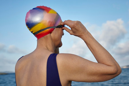 Mature woman wearing swimming costume and swimming hat, standing beside the sea, adjusting swimming goggles, rear viewの写真素材