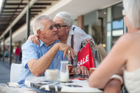 Woman shopper kissing senior man on cheek at sidewalk cafeの写真素材