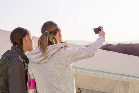 Two female friends outdoors, taking self portrait using smartphone, rear view