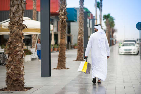 Rear view of man wearing dishdasha walking along street carrying shopping bags, Dubai, United Arab Emiratesの写真素材
