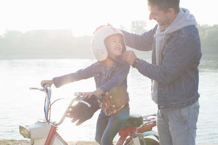 Young girl sitting on father's moped, father putting crash helmet on daughter's head, smilingの写真素材