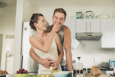 Romantic young woman feeding boyfriend whilst preparing breakfast at kitchen counter