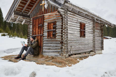 Young man reading map sitting outside log cabin in winter, Elmau, Bavaria, Germanyの写真素材