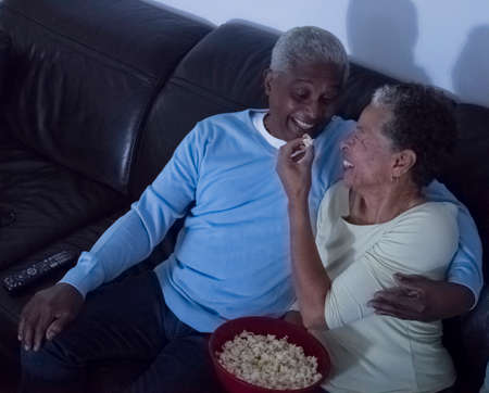 Senior couple sitting on sofa at night, woman feeding man popcornの写真素材