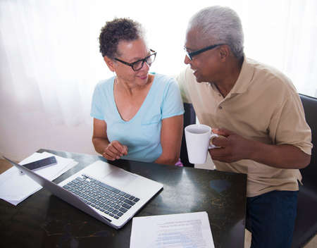 Senior couple sitting at table, using laptop, man holding coffee cupの写真素材
