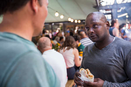 Male customers chatting and eating burgers at cooperative food market stall