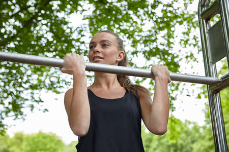 Young woman training in park, doing chin ups on exercise bars