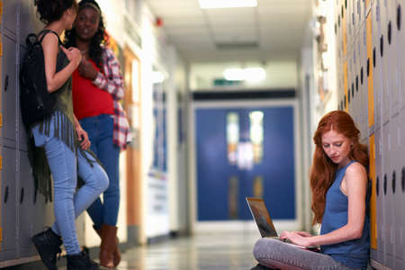 Young female college student sitting on locker room floor reading whilst typing on laptopの写真素材
