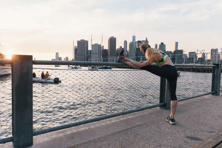 Young woman exercising outdoors, at waterfront, stretching leg, rear view, Brooklyn, New York, USA