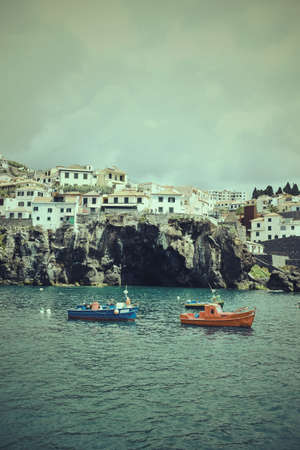 Boats in bay by clifftop buildings, Madeira, Camara de Lobos, Portugalの写真素材