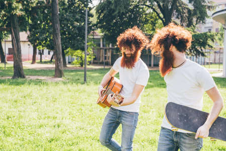 Young male hipster twins with red hair and beards strolling in park playing guitar