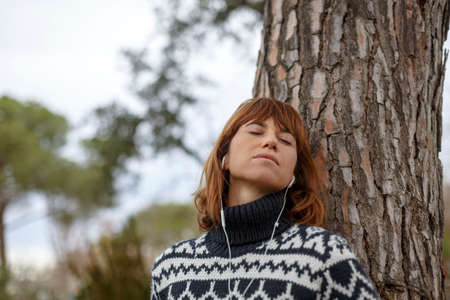 Woman leaning against tree wearing in ear headphones, eyes closedの写真素材