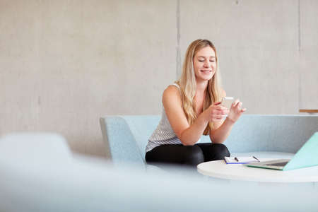 Young female student sitting on study space sofa reading smartphone texts at higher education...