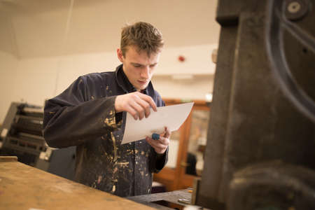 Young male printer inspecting paper for printing machinery in printing press workshop