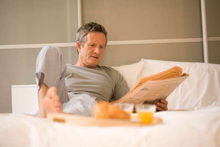 Senior man sitting up in bed reading newspaperの写真素材