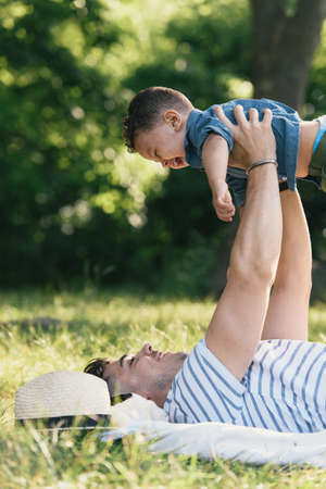 Young man lying on back holding up toddler son in Pelham Bay Park, Bronx, New York, USAの写真素材