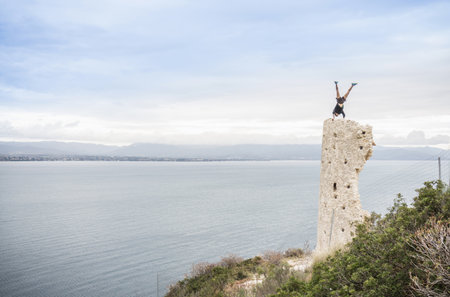 Male rock climber doing handstand on top of ruined tower on coast, Cagliari, Italyの写真素材