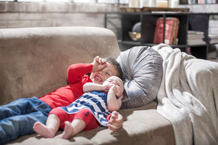 Boy and baby sister lying on sofa at homeの写真素材
