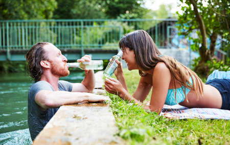 Young man in river, young woman lying on grass at edge of river, man drinking from beer bottleの写真素材