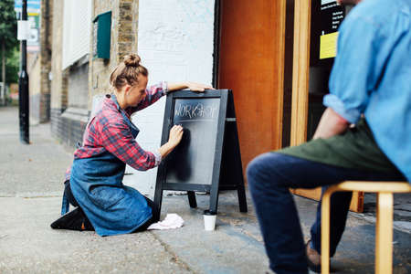 Young woman kneeling on pavement and writing on blackboard outside workshopの写真素材