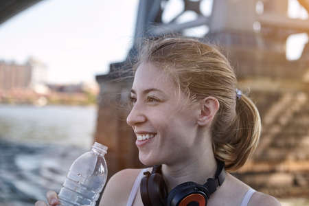 Young woman beside river, wearing headphones around neck, holding water bottle, New York City, USA